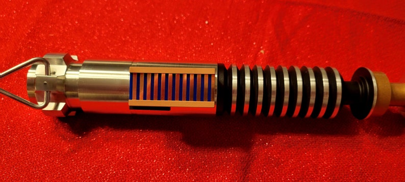May include: A detailed, metallic lightsaber prop with a silver handle, black and silver ribbed grip, and a section with blue and copper-colored details. The prop is set against a red background.