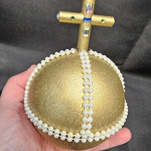 Könnte beinhalten: Goldfarbener, kugelartiger Schmuck mit einem Kreuz, verziert mit Kunstperlen und Edelsteinen. Das Kreuz hat grüne, blaue und irisierende Akzente. Der Schmuck wird in einer Hand vor dunklem Hintergrund gehalten.