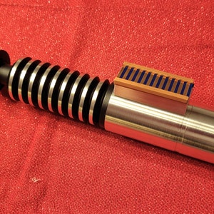 May include: A metallic lightsaber prop with silver, black, and gold accents. The hilt features a ribbed design and a rectangular detail with blue and gold stripes. The prop is set against a red background.