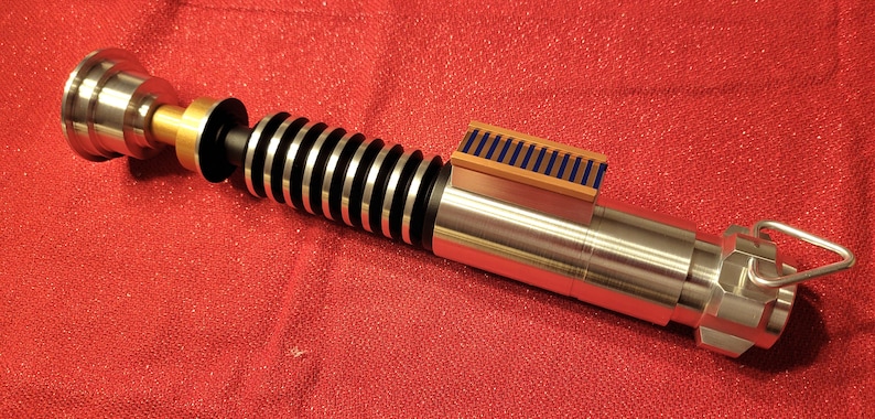 May include: A metallic lightsaber prop with silver, black, and gold accents. The hilt features a ribbed design and a rectangular detail with blue and gold stripes. The prop is set against a red background.