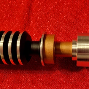 May include: A detailed view of a lightsaber hilt, featuring a black and silver ribbed grip, a gold-colored midsection, and a silver emitter. The lightsaber is set against a red background.