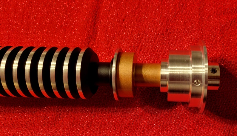 May include: A detailed view of a lightsaber hilt, featuring a black and silver ribbed grip, a gold-colored midsection, and a silver emitter. The lightsaber is set against a red background.