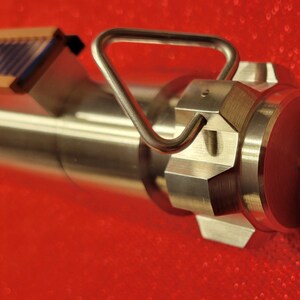 May include: Close-up of a metallic, industrial-looking device with a handle and a series of hexagonal and cylindrical components. The device has a handle and a small rectangular piece with blue and brown details. The background is a solid red.