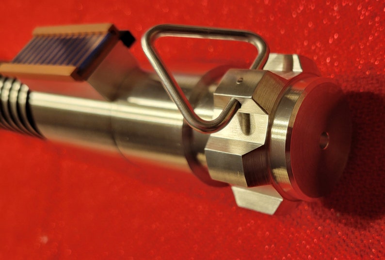 May include: Close-up of a metallic, industrial-looking device with a handle and a series of hexagonal and cylindrical components. The device has a handle and a small rectangular piece with blue and brown details. The background is a solid red.