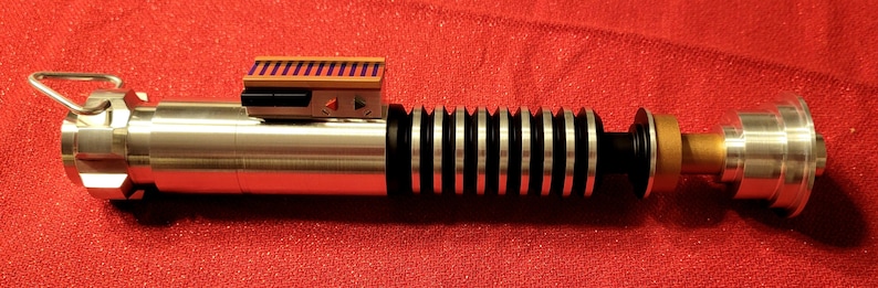 May include: A metallic lightsaber prop with silver, black, and gold accents. The hilt features a ribbed design, a rectangular control box with purple stripes, and a ring at the end. The prop is set against a red background.