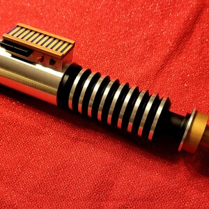 May include: A silver and black lightsaber prop with a gold and brown accent. The prop has a ribbed handle and a rectangular brown and black detail near the top. The lightsaber is set against a red background.