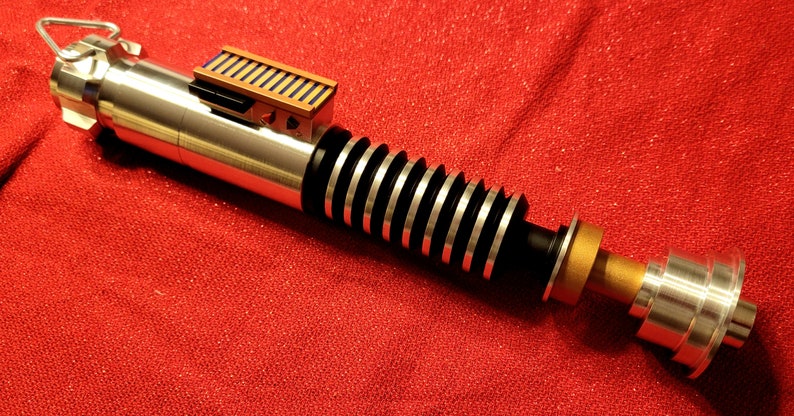 May include: A silver and black lightsaber prop with a gold and brown accent. The prop has a ribbed handle and a rectangular brown and black detail near the top. The lightsaber is set against a red background.