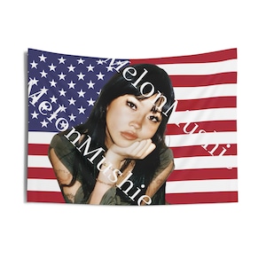 May include: A woman with long black hair is pictured in front of an American flag. The text "Melon Mushie" is written diagonally across the flag.