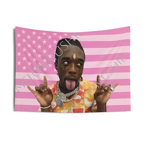 May include: A pink and white tapestry featuring a portrait of a person with their tongue out and hands in a rock and roll gesture. The background is a pink American flag design. The person is wearing jewelry and a patterned shirt.