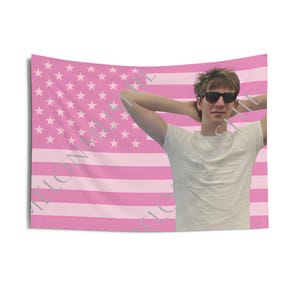May include: A pink American flag tapestry with white stars and pink stripes. A person wearing sunglasses and a white t-shirt is in front of the tapestry, with their arms behind their head.
