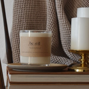 Puede incluir: Una vela blanca en un tarro de vidrio transparente con una etiqueta marrón que dice "be still" y una cita sobre el Señor que lucha por ti. La vela está sentada sobre una pila de libros con una manta marrón y otras dos velas en el fondo.