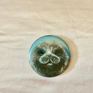 May include: A round button featuring a painted seal with a light blue background. The seal has dark eyes, a dark nose, and white accents. The button is a cute, whimsical design.