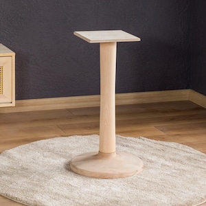 May include: A light-colored wooden pedestal table with a square top, a cylindrical stem, and a circular base. The table stands on a beige, round rug, with a wooden cabinet and a framed artwork in the background.
