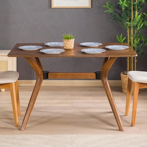 Puede incluir: Una mesa de comedor de madera con una tapa marrón y patas de metal negro. La mesa está puesta con cuatro platos blancos con diseños florales azules y una pequeña planta en maceta en el centro. Dos sillas de madera con cojines beige y respaldos de caña trenzada se colocan a cada lado de la mesa. Una planta en maceta con hojas verdes está en el fondo.