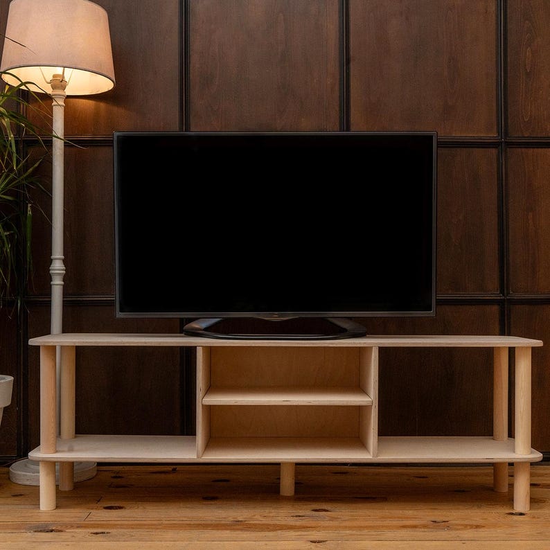 May include: A light-colored wooden TV stand with a flat-screen TV on top. The stand has two open shelves and a central compartment with two shelves. A floor lamp with a beige shade stands to the left.