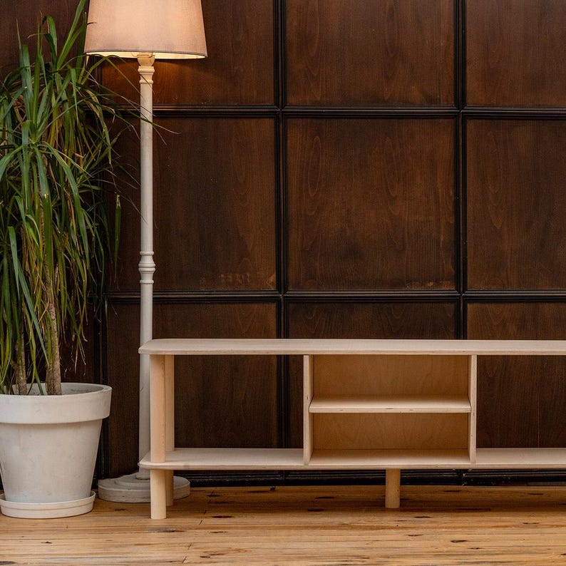 May include: A light-colored wooden media console with open shelving and a simple design. The console is set against a dark wood paneled wall. A tall, off-white floor lamp with a beige lampshade stands to the left.