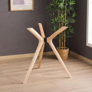 Puede incluir: Una base de mesa de madera clara con cuatro patas anguladas que se extienden hacia afuera desde un conector circular central de color negro. La estructura está diseñada para soportar un tablero, mostrando un acabado de veta de madera natural.