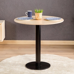 May include: A round, light wood table with a black metal base. Two blue and white patterned plates, a mug, and a small plant in a woven basket sit on the table. The table is approximately 30 inches in diameter.
