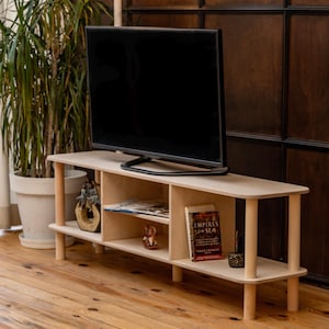 May include: A light-colored wooden TV stand with a flat-screen television on top. The stand has two shelves and cylindrical legs. Books and decorative items are visible on the shelves. A potted plant is in the background.