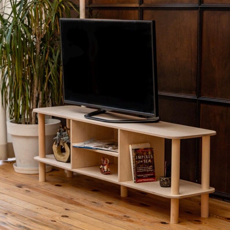 May include: A light-colored wooden TV stand with a flat-screen television on top. The stand has two shelves and cylindrical legs. Books and decorative items are visible on the shelves. A potted plant is in the background.