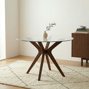 Può includere: Un tavolo da pranzo rotondo con piano in vetro trasparente e base in legno marrone scuro con quattro gambe angolate che si incontrano in un punto centrale. Un piccolo vaso in ceramica chiara con un ramo di foglie poggia sul vetro.