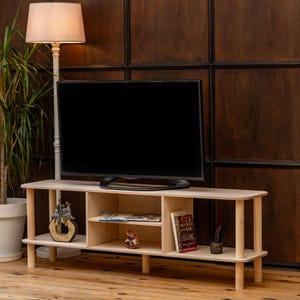 May include: A light-colored wooden TV stand with a flat-screen television on top. The stand has open shelves with decorative items and books. A floor lamp and a potted plant are in the background.