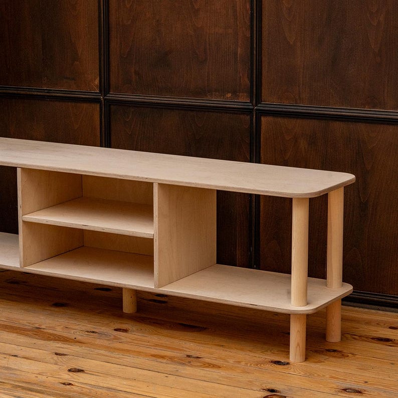 May include: A light-colored wooden console table with a rectangular top and rounded edges. The table has two shelves and is supported by cylindrical legs. The wood grain is visible, and the piece has a minimalist design.