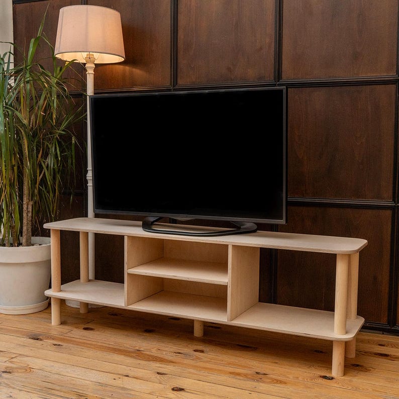 May include: A light-colored wooden TV stand with a rectangular shape and rounded edges. The stand has two shelves and is supported by cylindrical legs. A large black television sits on top of the stand. A floor lamp and a potted plant are in the background.