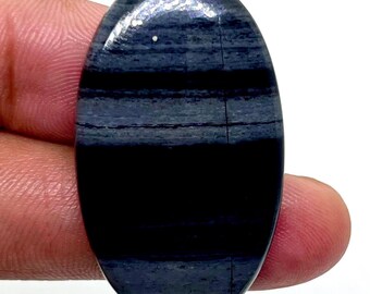 May include: An oval-shaped gemstone with a polished surface. The stone features a layered pattern of dark gray and black bands. A thin, vertical crack runs through the center of the stone. The gemstone is held between fingers.