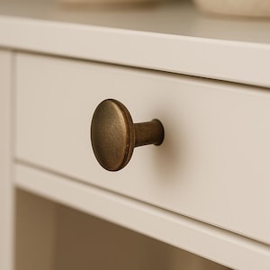 Könnte beinhalten: Nahaufnahme einer weißen Schublade mit einem ovalen Knopf in Bronzefarbe. Der Knopf ist mittig auf der Schublade angebracht, die Teil eines weißen Schranks ist. Im Hintergrund ist eine verschwommene Pflanze in einem beigen Topf zu sehen.
