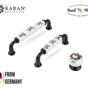 Könnte beinhalten: Zwei weiße Keramik-Schrankknöpfe mit schwarzen Blumenmustern und schwarzen Metallsockeln. Ein einzelner Knopf ist ebenfalls im Bild zu sehen. All Musterarbeiten an Hand Made Produkten sind handgefertigt.