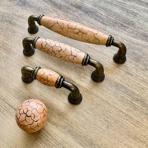 Puede incluir: Tres pomos de armario de bronce antiguo con un acabado de cerámica beige agrietada. Los pomos son de diferentes tamaños, siendo el más pequeño un pomo redondo.