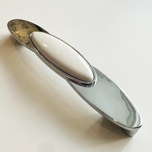 Può includere: Un pomello per armadio cromato con un inserto ovale in ceramica bianca. Il pomello ha una finitura liscia e lucida.