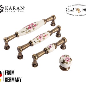 Könnte beinhalten: Satz von drei antiken goldenen Schrankknöpfen mit weißen Keramiksockeln und rosafarbenen Blumendesigns. Die Knöpfe sind in einer Reihe vom größten zum kleinsten angeordnet. Der kleinste Knopf ist ein runder Knopf. Der Text "Hand Made" ist im Hintergrund sichtbar.