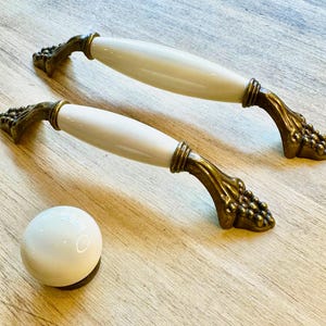 Könnte beinhalten: Zwei weiße Keramik-Schrankknöpfe mit verzierten goldenen Metallsockeln. Die Knöpfe haben ein Rankenmuster.