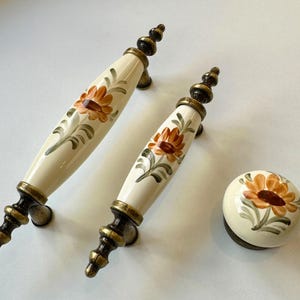 Può includere: Tre tiretti per cassetti in ceramica con un design floreale. I tiretti sono dipinti con uno sfondo bianco e presentano fiori arancioni e marroni. I tiretti hanno una finitura in ottone.