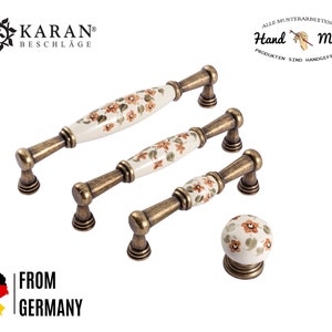 Puede incluir: Conjunto de tres pomos de armario de oro antiguo con manijas de cerámica blanca con un diseño floral en tonos naranja y verde. Los pomos están hechos en Alemania. "Hand Made" y "From Germany" también son visibles en la imagen.