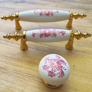 Pode incluir: Duas puxadores de gaveta de cerâmica branca com detalhes dourados e desenhos florais rosa. Uma maçaneta de cerâmica branca combinando com detalhes dourados e desenhos florais rosa.