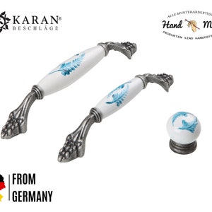 Pode incluir: Dois botões de armário de cerâmica branca com desenhos florais azuis e bases de metal prateado. Os botões são mostrados contra um fundo branco. O texto "Hand Made" é visível no canto superior direito da imagem.