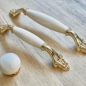 Könnte beinhalten: Zwei weiße Keramik-Schrankknöpfe mit goldenen Akzenten und ein einzelner weißer Keramik-Knopf mit goldener Basis.