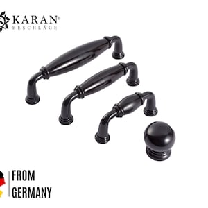 Pode incluir: Quatro botões e puxadores de armário pretos em vários tamanhos. Os botões e puxadores têm uma forma oval lisa, ligeiramente arredondada. KARAN® BESCHLAGE está impresso no topo da imagem.