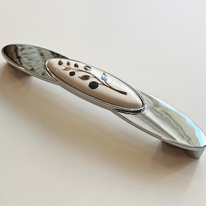 Könnte beinhalten: Ein Chrom-Schrankgriff mit einem weißen Keramikeinsatz, der mit einem Blumendesign verziert ist. Das Blumendesign ist in Gold-, Schwarz- und Blautönen gehalten.