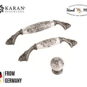 Pode incluir: Conjunto de três botões de armário de cerâmica branca com acabamento prateado e um padrão preto em espiral. Os botões são etiquetados como "Hand Made" e "Karan Beschläge".