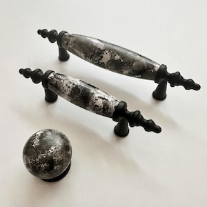 Könnte beinhalten: Drei schwarze und weiße Keramik-Schrankknöpfe mit Marmormuster. Die Knöpfe haben eine schwarze Metallbasis mit einem dekorativen Scroll-Design.
