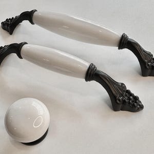 Könnte beinhalten: Zwei weiße Keramik-Schrankknöpfe mit schwarzen Metallsockeln in Form von Trauben. Die Knöpfe haben eine glatte, matte Oberfläche. Es gibt auch einen einzelnen weißen Keramik-Knopf mit einem schwarzen Metallsockel.