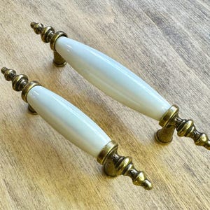 Può includere: Due maniglie per mobili con manici in ceramica bianca e dettagli in metallo color ottone antico. Le maniglie hanno un design classico, con un centro cilindrico e terminali decorativi. Sono poste su uno sfondo chiaro con venature del legno.