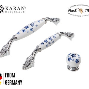 Peut inclure: Deux boutons de cabinet en céramique blanche avec des motifs floraux bleus et des bases en métal argenté. Un seul bouton est visible en bas au centre de l'image. Le texte "Hand Made" est visible dans le coin supérieur droit de l'image.