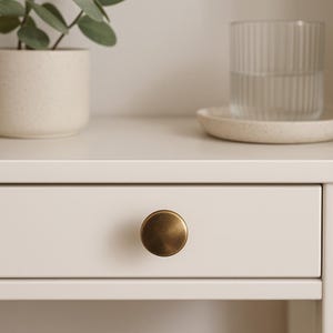 May include: Close-up of a white drawer with a round, antique brass knob. The drawer is part of a white cabinet or desk. A small potted plant, a glass of water, and a small plate are on the surface above the drawer.