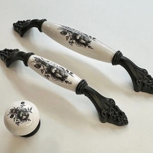 Könnte beinhalten: Zwei schwarze und weiße Keramik-Schubladengriffe mit Blumendesigns und schwarzen Metallsockeln. Ein passender schwarzer und weißer Keramik-Knopf mit Blumendesign ist ebenfalls enthalten.