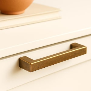 Può includere: Primo piano di una maniglia per cassetto rettangolare color oro. L'hardware è montato su un mobile bianco. Una pila di libri e una ciotola di terracotta sono visibili sullo sfondo.